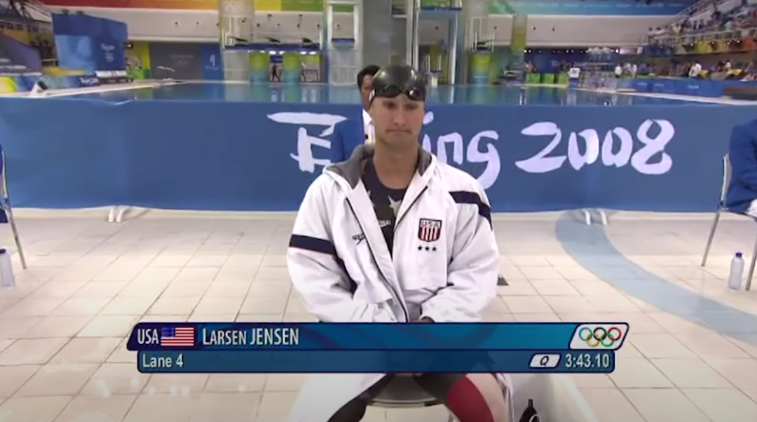 A male swimmer in a USA team jacket and swim goggles sits poolside at the 2008 Beijing Olympics. The screen displays "Larsen Jensen," "Lane 4," and a time of 3:43.10. The Olympic logo and pool are visible in the background.