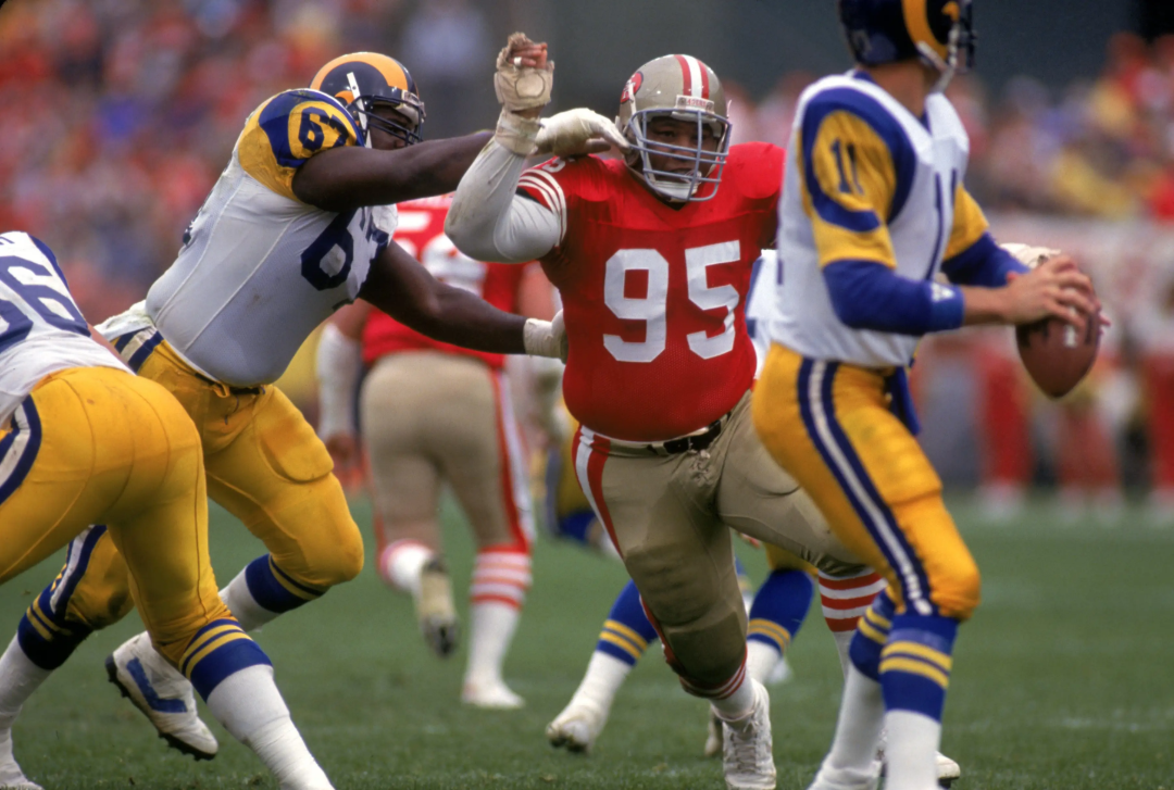 A football player in a red jersey (number 95) rushes past a blocker in a white and yellow uniform, while another player in a blue, yellow, and white uniform prepares to throw the ball during a game.