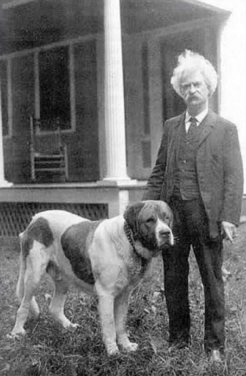 A man with white hair and a mustache stands next to a large St. Bernard dog in front of a porch with columns. Both are looking toward the camera. The background shows a house with a wooden rocking chair.