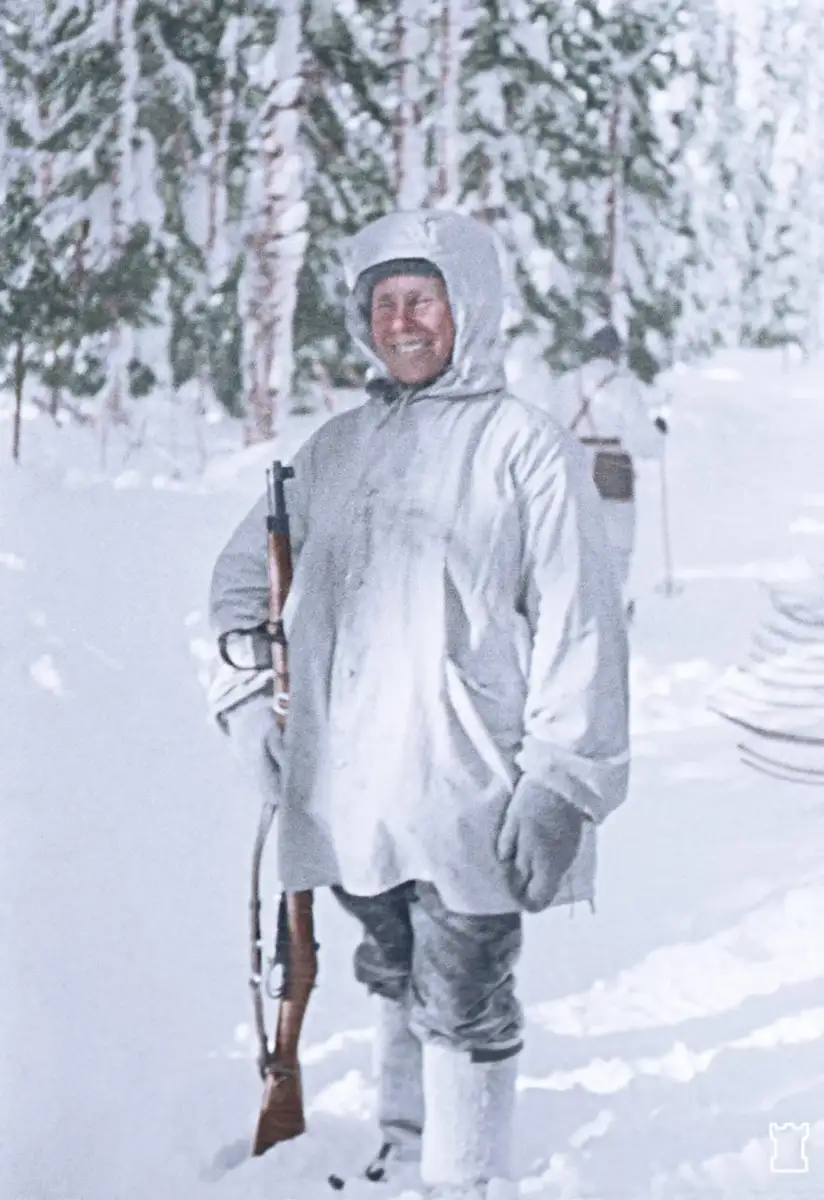 A person in white winter camouflage stands smiling in a snowy forest, holding a rifle. They are dressed in warm clothing and boots, with snow and trees in the background.