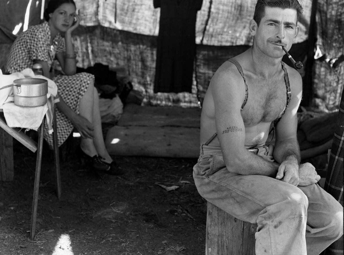 A shirtless man with a pipe sits on a wooden bench inside a tent, while a woman in a checkered dress sits nearby, resting her face on her hand. Household items are visible on a small table to the left.