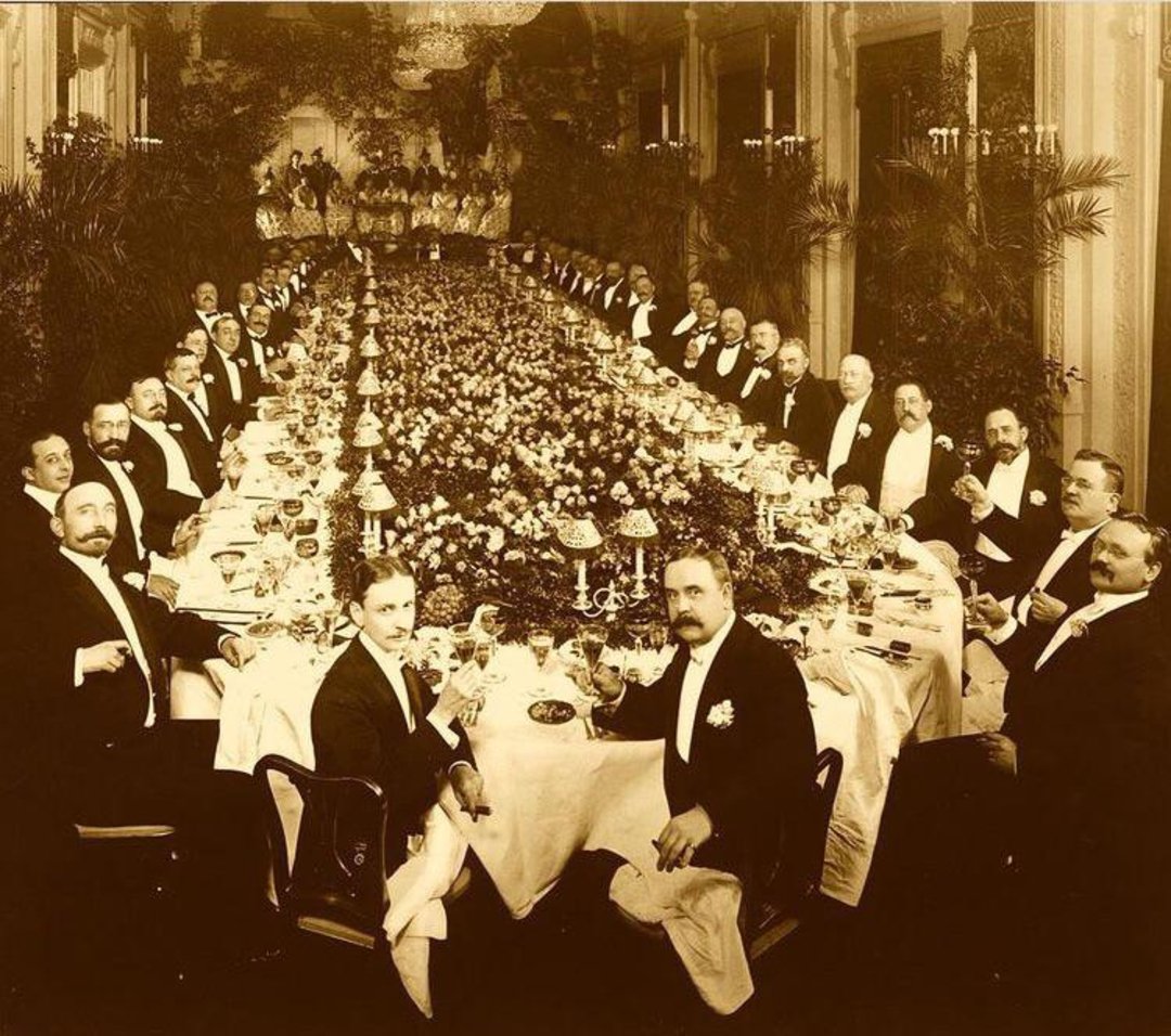A large group of men in formal attire sit around a long, ornate dining table covered with elaborate floral arrangements and glassware in an elegant, high-ceilinged room, raising glasses in a toast.