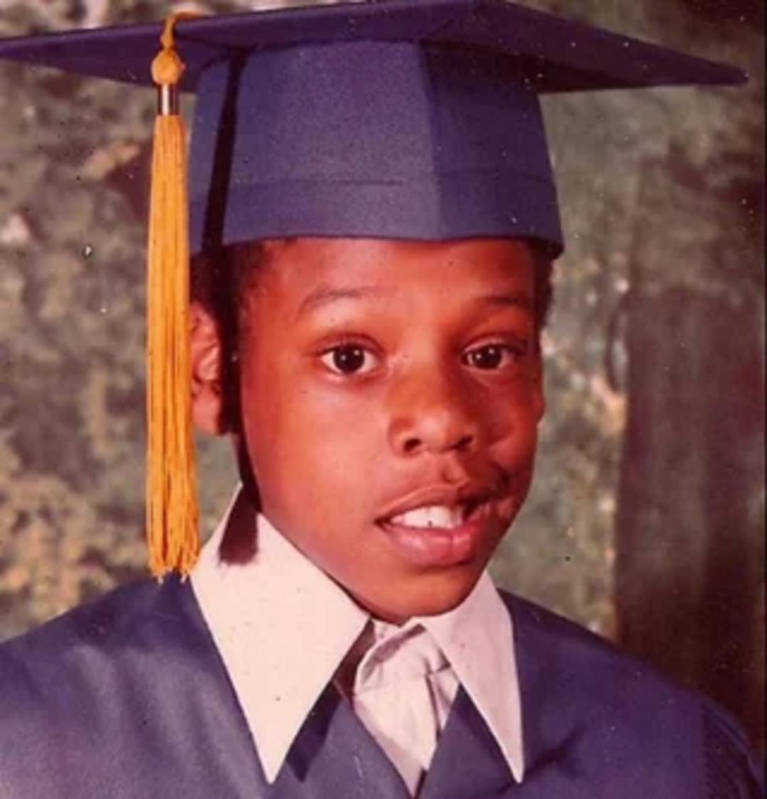 A young Black boy wearing a navy blue graduation cap and gown with a yellow tassel stands in front of a mottled green background, looking at the camera and smiling slightly.