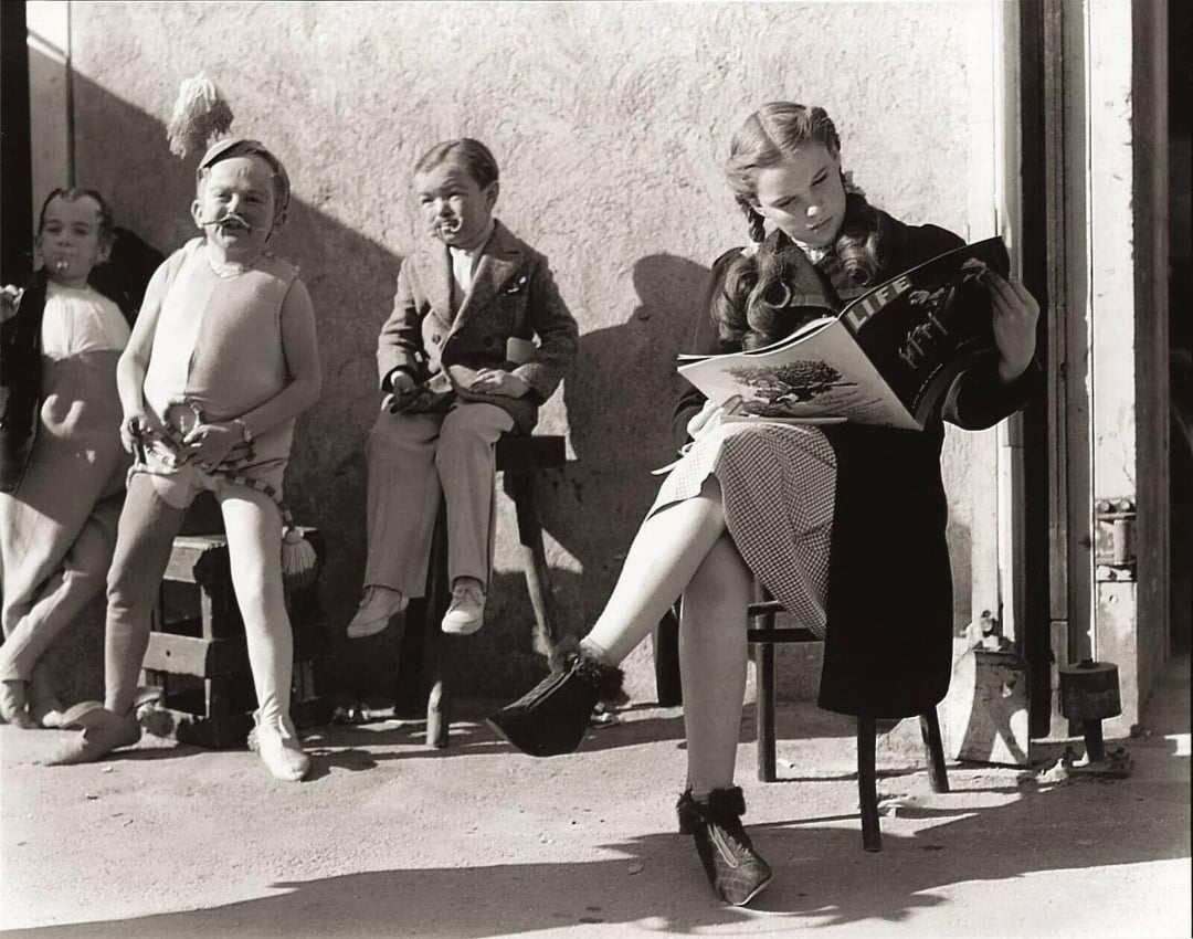 A young woman sits on a chair reading a LIFE magazine outside, while three people in costume, likely actors, stand or sit nearby against a textured wall in sunlight.