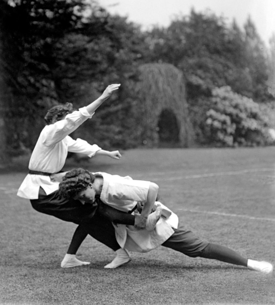 Two women in early 20th-century attire practice jujutsu outdoors on grass, with one woman throwing the other over her hip. Trees and shrubs are visible in the background.