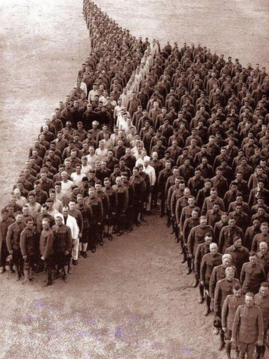 A large group of uniformed soldiers stands arranged in formation to create the shape of a human face in profile, viewed from above on an open, sandy ground. The photo is in black and white.