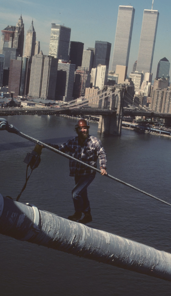 A construction worker in a plaid shirt and hard hat stands on a large suspension cable above the East River, with the Brooklyn Bridge and the Twin Towers of the World Trade Center in the background.