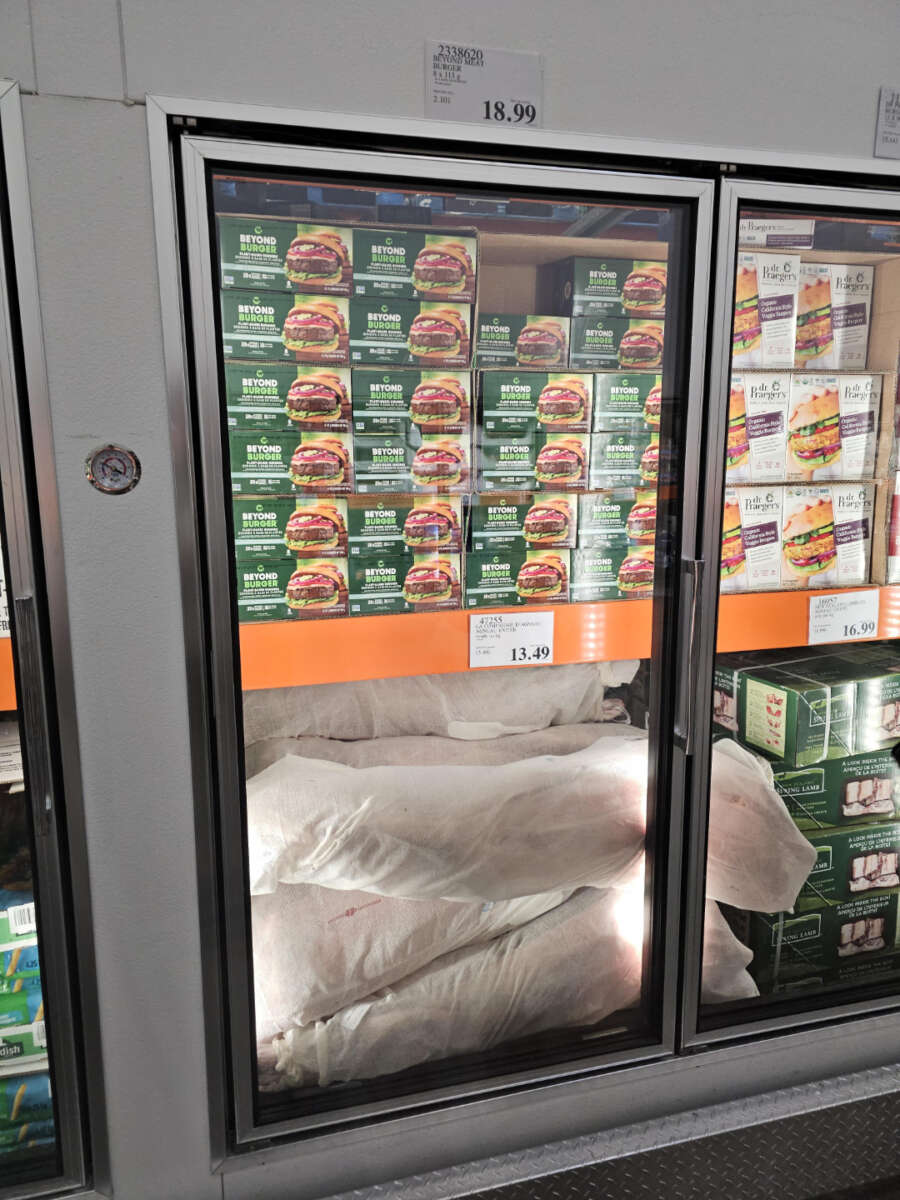 A freezer section displays stacked boxes of Beyond Burger plant-based patties on the top shelf and large white bags filled with items on the bottom shelf. Price labels are visible on the glass door.