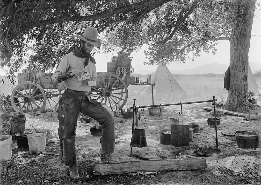 A cowboy stands at a rustic outdoor camp, pouring coffee into a cup. Cooking pots hang over a fire, with supplies, a wagon, and a tent in the background beneath shady trees.