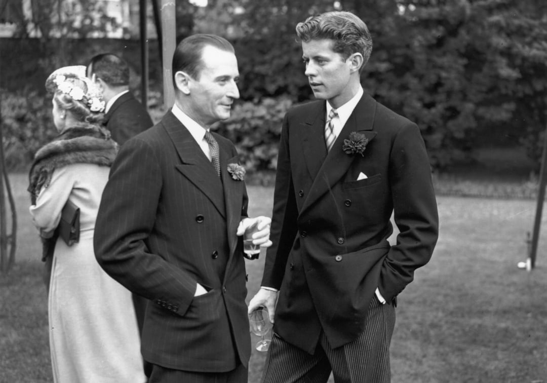 Two men in suits converse outdoors at a formal event, one holding a drink. In the background, a woman in a coat with a fur collar and another man are also visible. Lush trees and grass complete the setting.