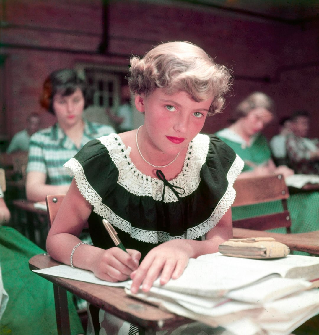 A young girl with short blonde hair, wearing a black dress with a white lace collar, sits at a desk writing in a notebook in a classroom with other students in the background.