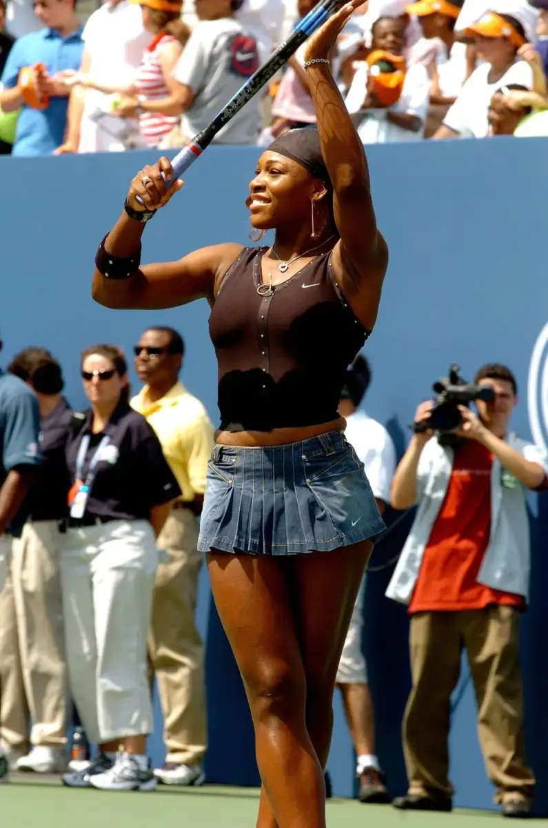 A female tennis player celebrates on the court, holding her racket overhead, smiling, and wearing a black sleeveless top and blue pleated skirt. Spectators and photographers are visible in the background.
