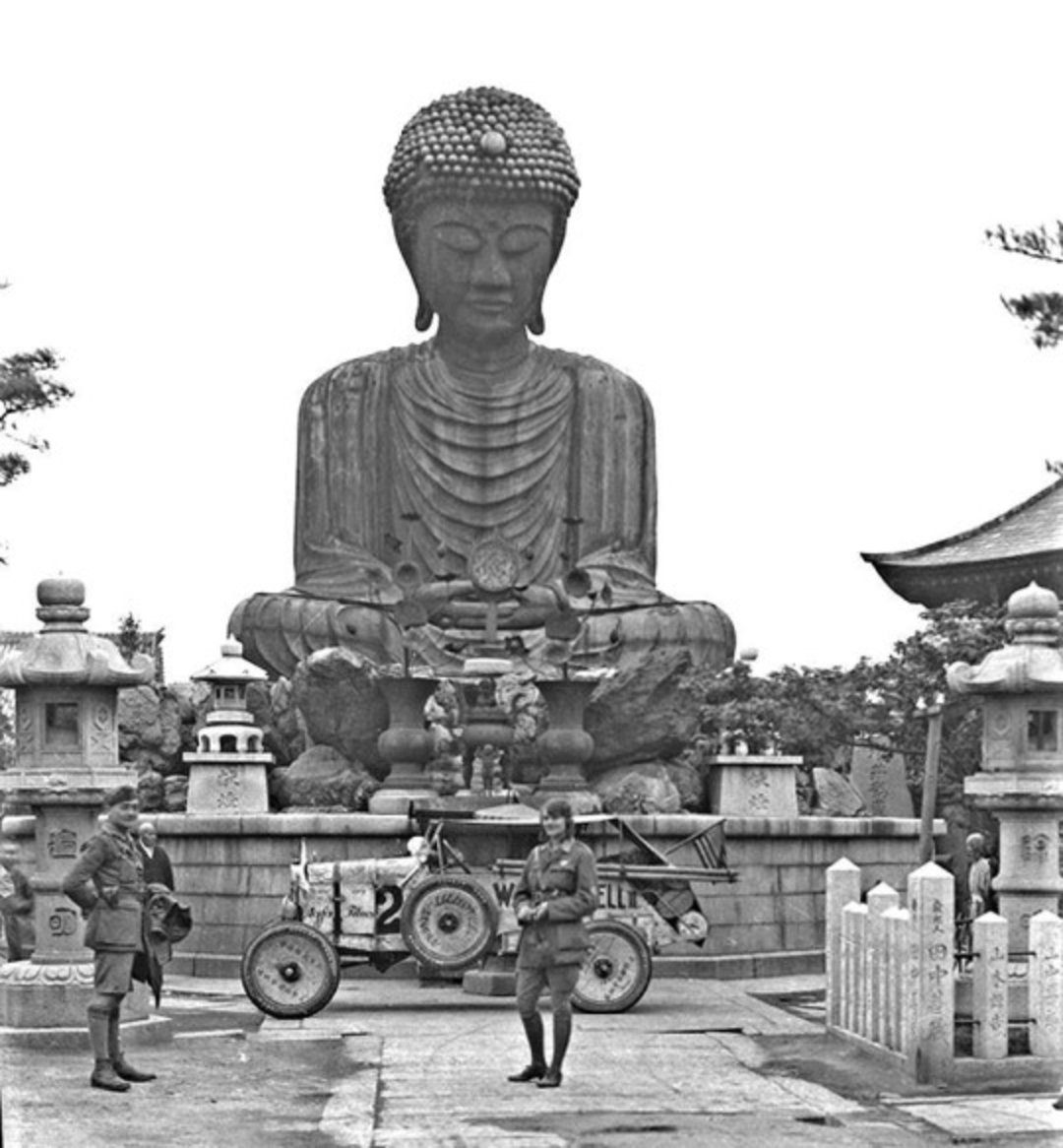 A black and white photo of a large seated Buddha statue outdoors, with three soldiers standing in front and a vintage race car parked nearby. Traditional Japanese lanterns and temple architecture are also visible.