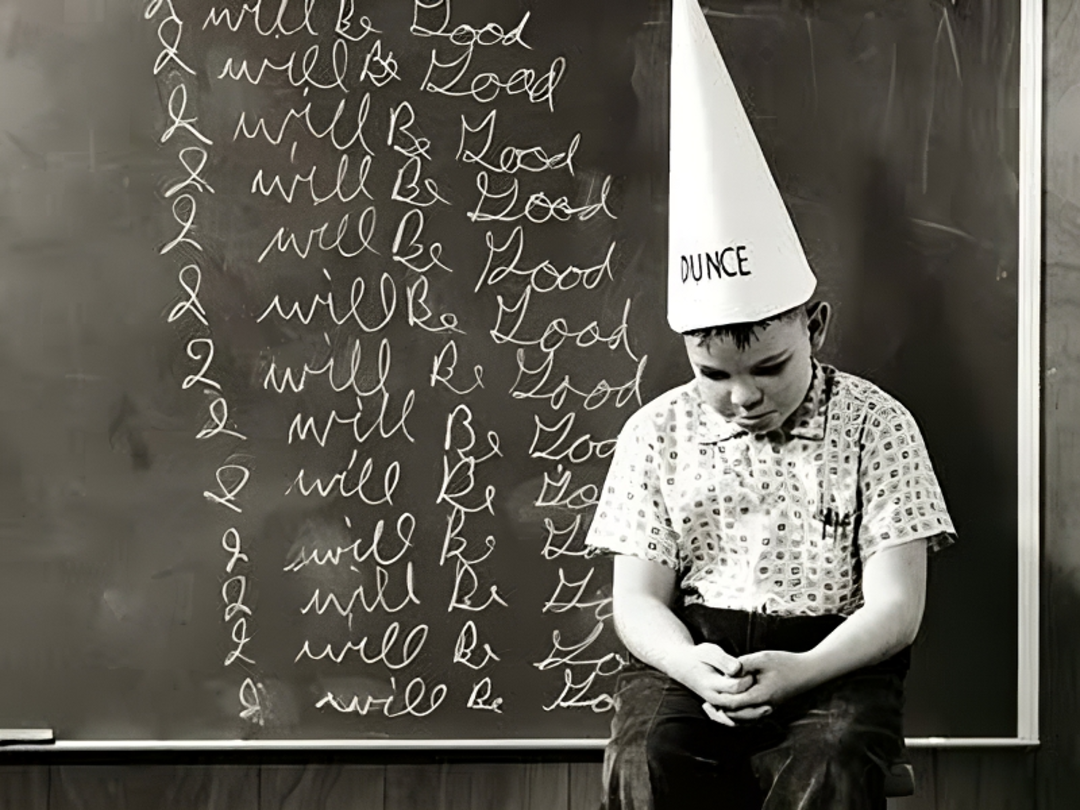 A boy sits on a stool wearing a dunce cap, looking down sadly. Behind him, a chalkboard displays repeated lines of "I will be good" written in cursive.