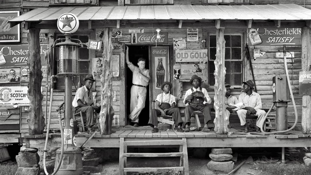 Black-and-white photo of six people sitting and standing on the porch of a rustic wooden store, surrounded by vintage advertisements and an old gas pump. The mood is relaxed and the building appears weathered.