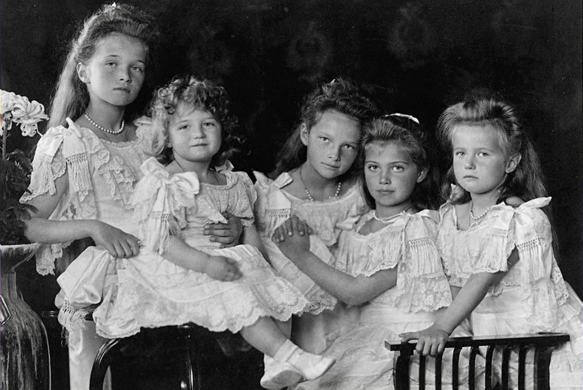 Five young girls wearing white lace dresses sit and pose together for a formal portrait against a dark background. They have serious expressions and are seated closely, with flowers visible on the left.