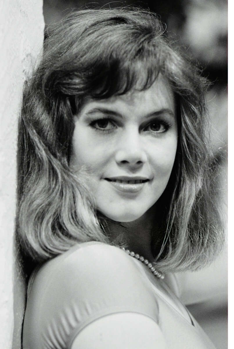 A woman with shoulder-length hair wearing a necklace and a light top smiles softly while leaning against a white wall, in a black and white portrait.