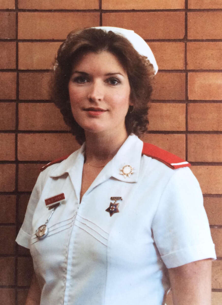 A woman wearing a white nurse uniform with red epaulettes, a white cap, and multiple pins stands in front of a brick wall, looking at the camera and smiling slightly.