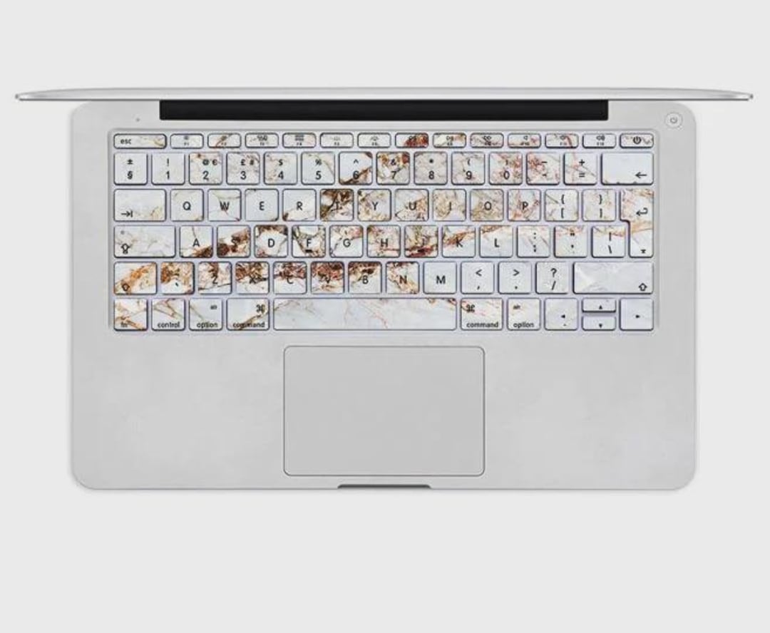 A silver laptop viewed from above, with a marble-patterned keyboard cover featuring brown, tan, and gold accents on the keys. The trackpad and the rest of the laptop appear clean and minimalistic.