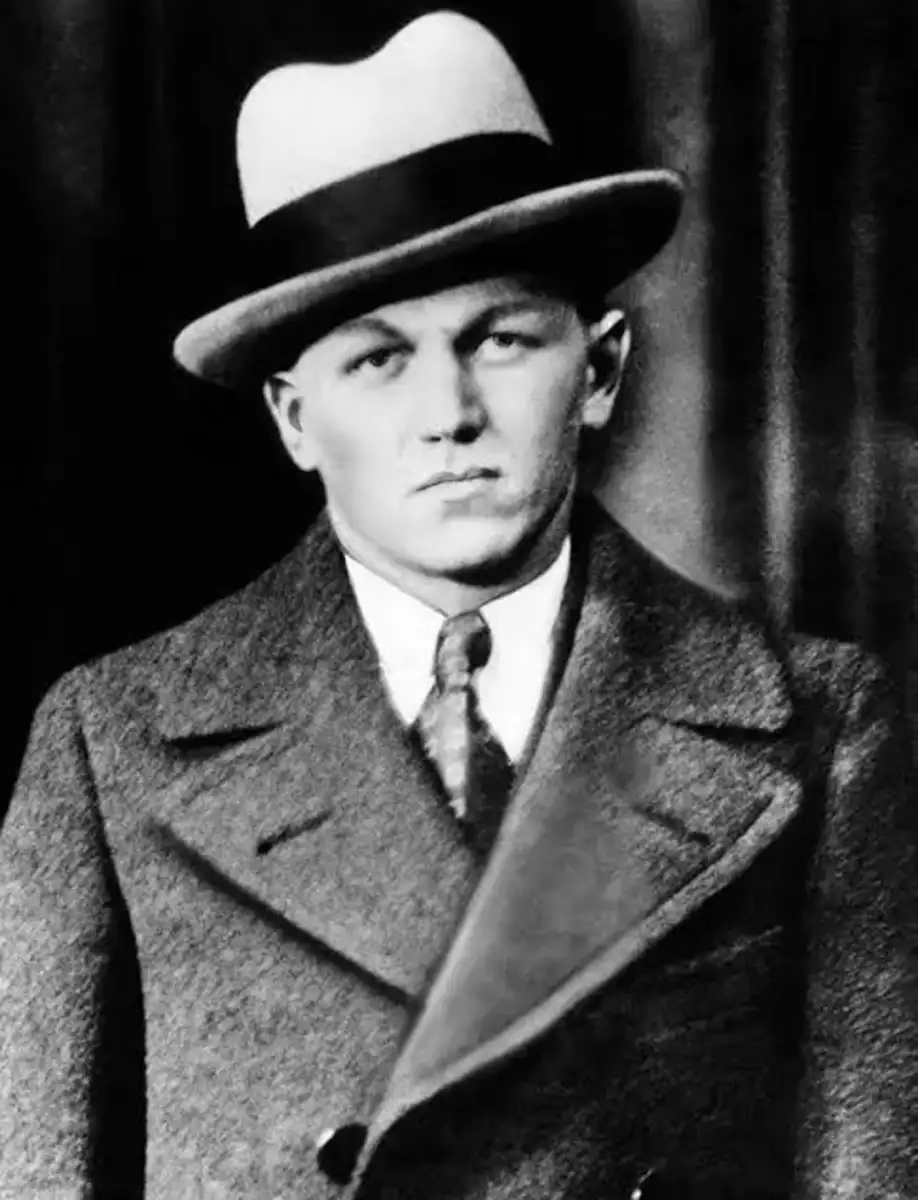 Black-and-white photo of a stern-looking man wearing a light-colored fedora hat, a double-breasted overcoat, a shirt, and a tie, posing in front of a dark background.