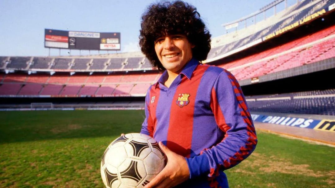 A smiling soccer player with curly hair wearing a blue and red FC Barcelona jersey stands on a stadium field holding a soccer ball. The stadium seats are mostly empty in the background.