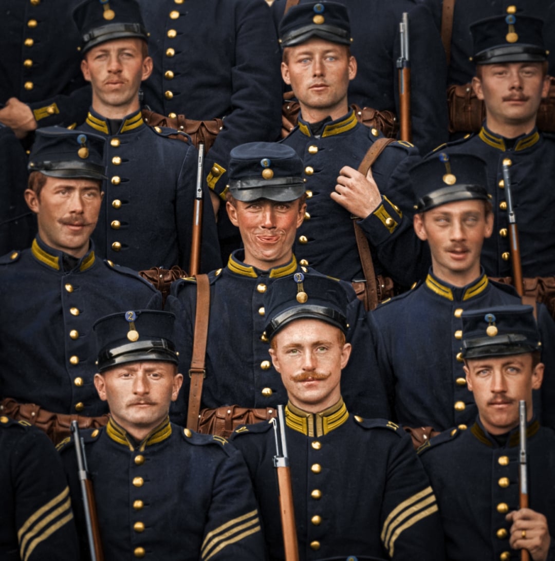 A group of young men in dark blue military uniforms with gold buttons and trim pose for a formal photograph, each holding a rifle and wearing matching caps. Most have serious expressions and neat mustaches.