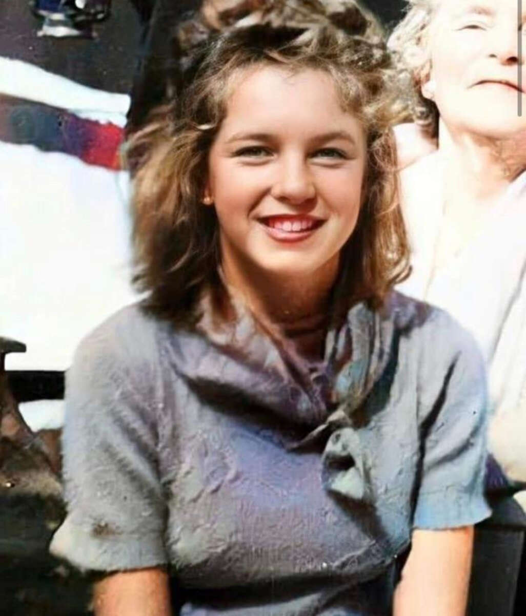 A young woman with wavy light brown hair and a big smile sits in front of an older woman. She wears a short-sleeved, textured blue dress with a scarf at the collar. The background is slightly blurred.