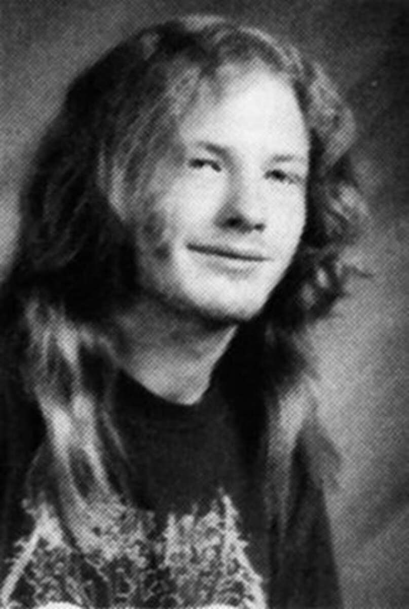 A black and white portrait of a young person with long, wavy hair wearing a dark graphic T-shirt, smiling slightly, and facing the camera. The background is plain and softly blurred.