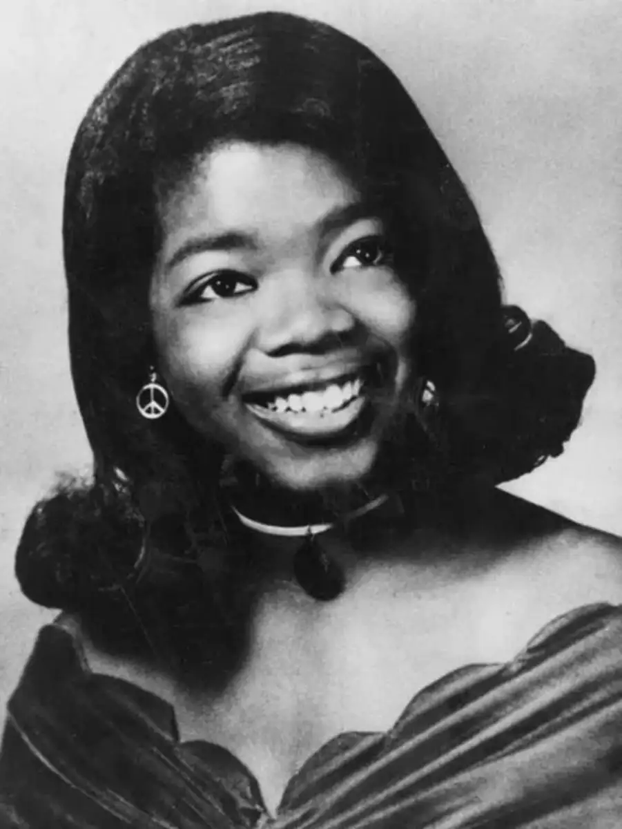 A young Black woman smiles in a black-and-white portrait, wearing a velvet off-the-shoulder gown, a choker necklace, and earrings with a peace symbol. Her hair is styled in soft waves.