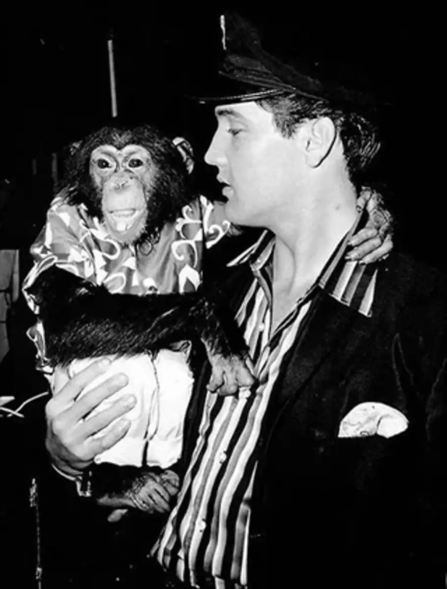 A man in a striped shirt and cap holds a chimpanzee dressed in a patterned shirt and pants. The photo is in black and white.