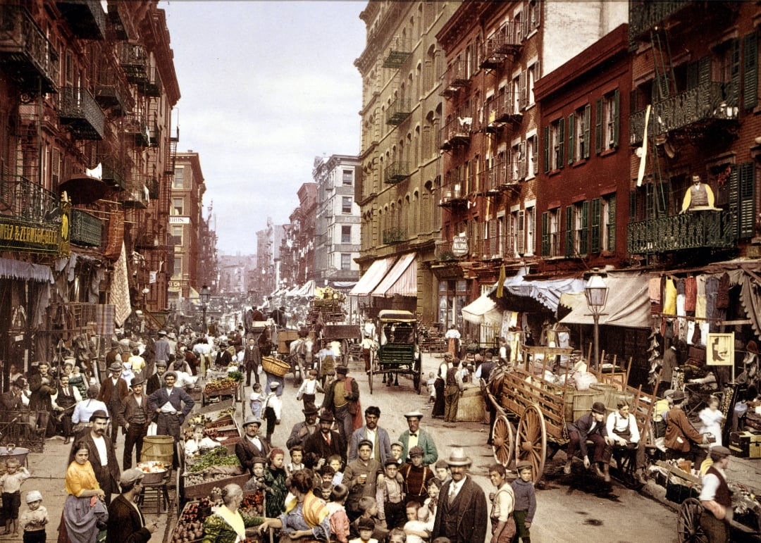 A bustling city street scene from the early 1900s with crowded sidewalks, market stalls, horse-drawn carts, and many people in period clothing surrounded by tall brick apartment buildings.