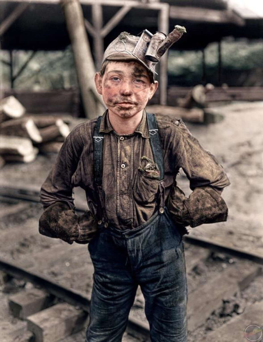 A young coal miner stands on a dirt path beside railway tracks, wearing dirty work clothes, suspenders, gloves, and a mining helmet with a light. His face and hands are covered in coal dust.
