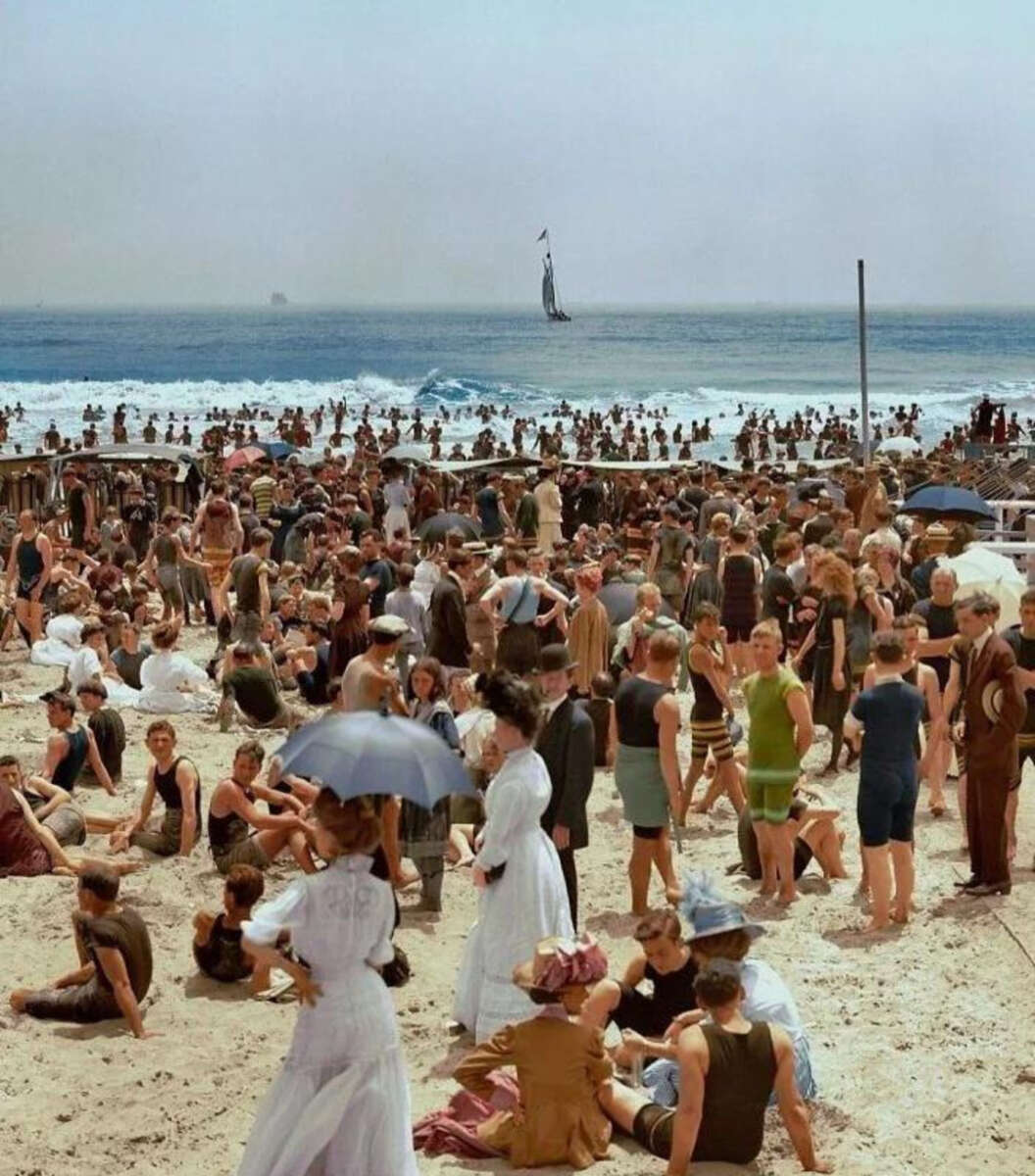 A crowded beach scene from the early 1900s, with people in vintage bathing suits and dresses, some sitting, some standing. A sailboat is visible on the ocean, and the sky is clear and blue.