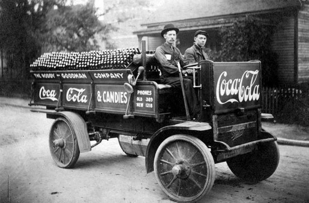Black and white photo of two men sitting on an old Coca-Cola delivery truck loaded with glass bottles. The truck has large wheels and advertisements for Coca-Cola and candies on its sides. A house is visible in the background.