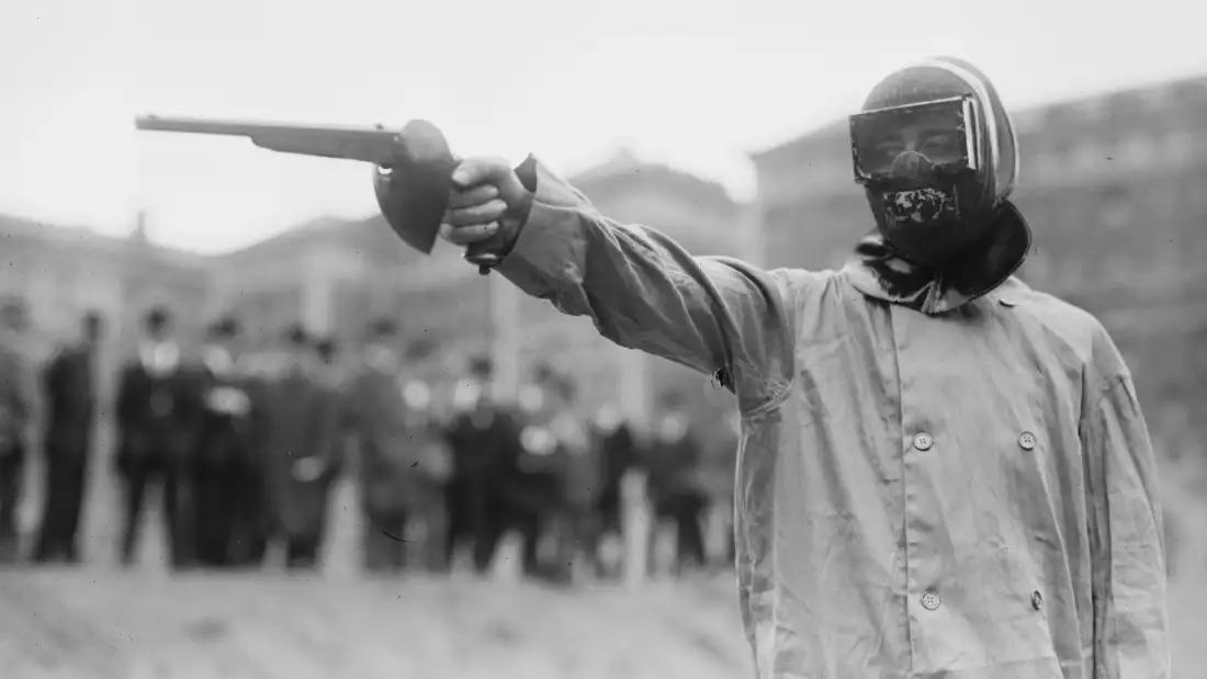 A person wearing a helmet, goggles, and a mask aims a pistol while standing outdoors; blurred group of people in the background. The image is black and white.
