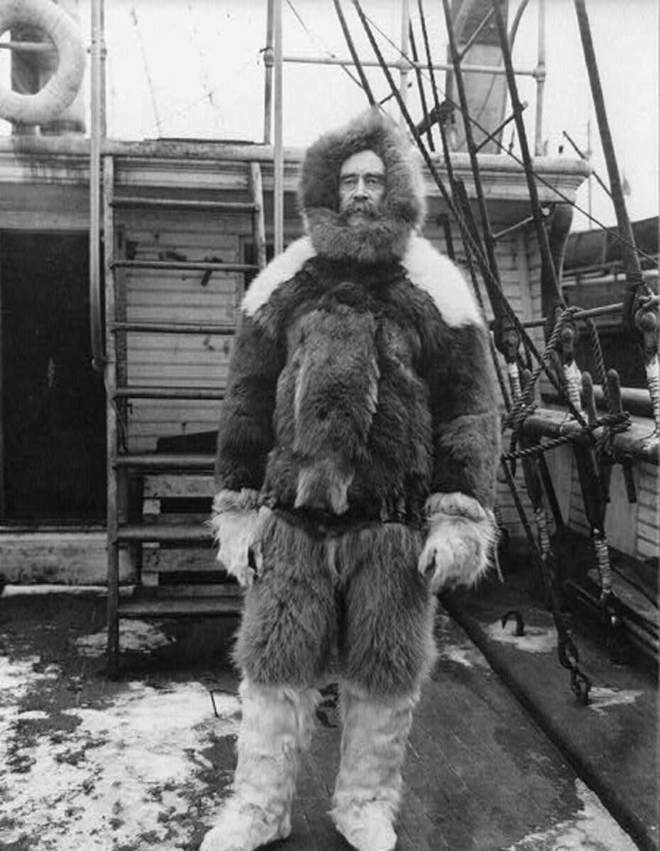 A person wearing a thick fur parka and pants stands on a wooden ship deck surrounded by ropes and snow, with a wooden structure and steps in the background.