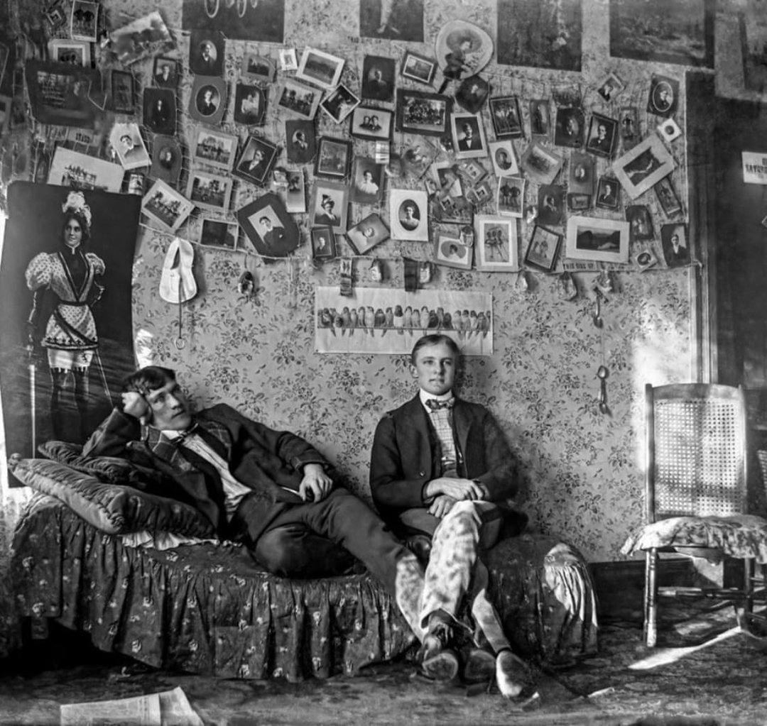 Two young men sit in a cluttered room; one reclines on a bed while the other sits on a chair. The walls behind them are covered with numerous photographs and portraits, creating a dense collage above patterned wallpaper.