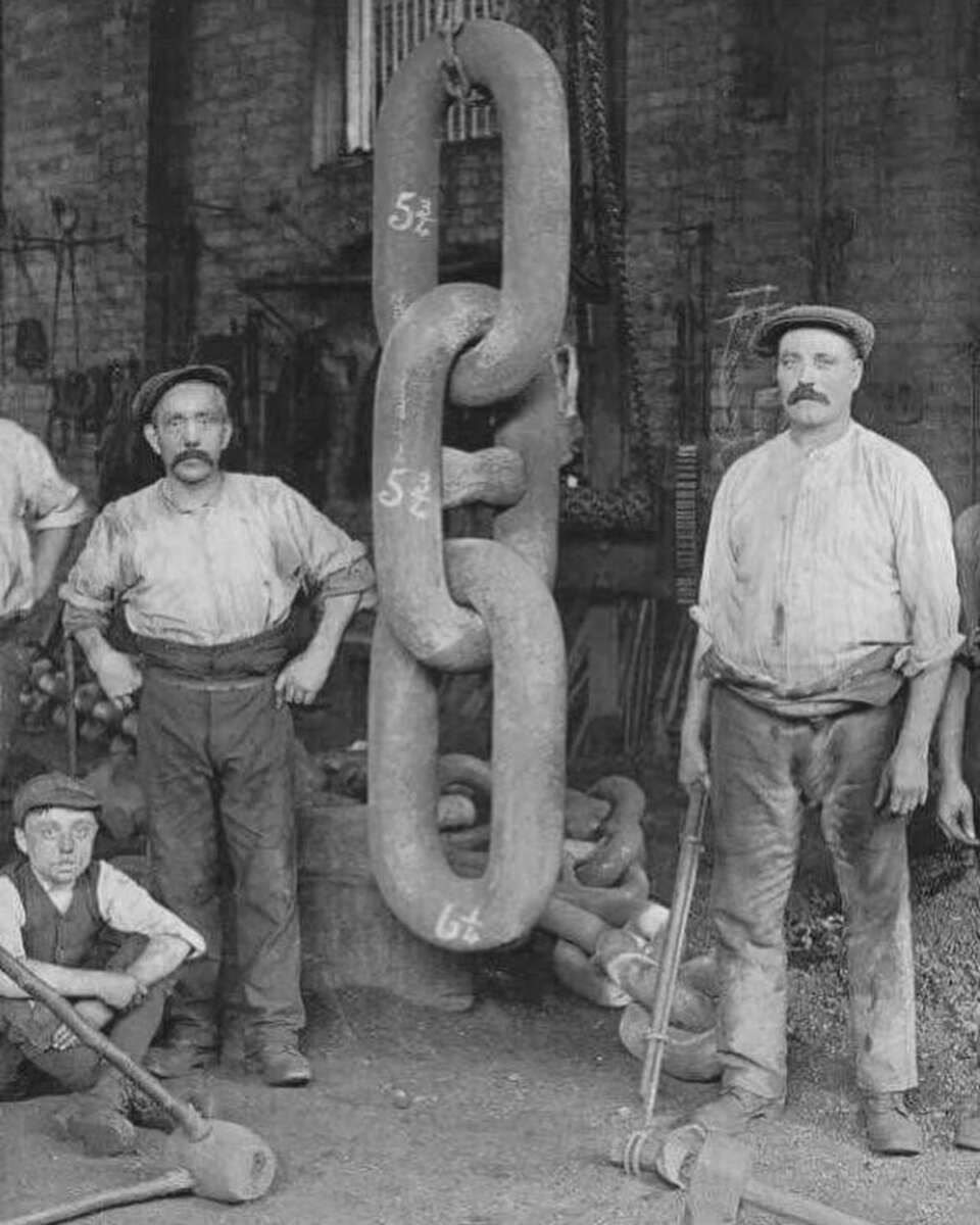 Two men in work clothes and flat caps stand beside a massive iron chain link, marked with measurements, in an old industrial workshop. A boy crouches nearby, and large tools and machinery are visible in the background.