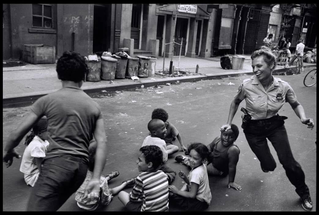 A smiling police officer chases laughing children sitting and playing in the middle of a city street lined with buildings, trash cans, and scattered debris.