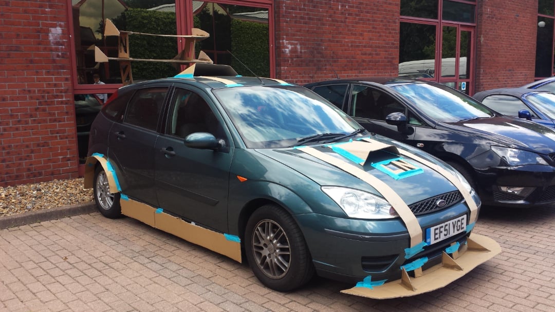 A dark green car covered with cardboard pieces taped on the hood, roof, sides, and wheels, parked outside a brick building next to another car. The cardboard is secured with blue tape in various shapes.