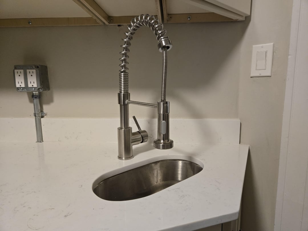 A modern stainless steel kitchen faucet with a flexible spring spout is installed above a small, oval-shaped sink on a white countertop. Electrical outlets and a light switch are visible on the wall.