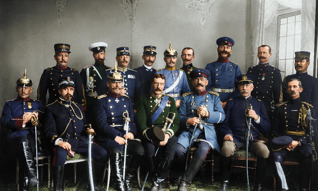 A group of men in ornate military uniforms from various countries, seated and standing in two rows, pose for a formal photograph in an ornately decorated room with a window on the right.