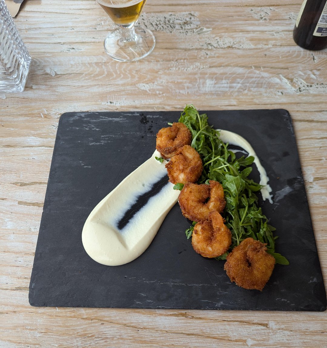 Three breaded and fried shrimp are served on a bed of arugula with a smear of creamy white sauce and a dark streak on a black slate plate, with a glass of beer and a bottle in the background on a wooden table.