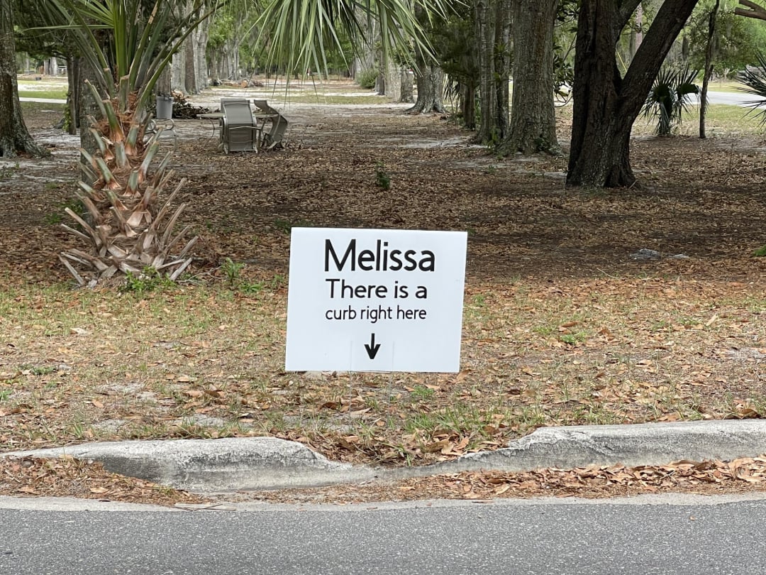 A white sign by the road reads, "Melissa There is a curb right here," with an arrow pointing down to a curb in front of it. Trees and outdoor chairs are visible in the background.