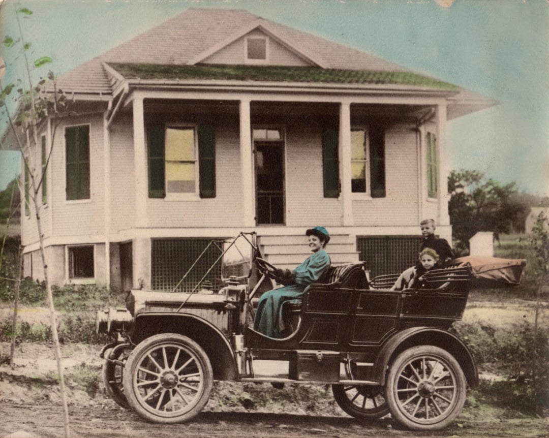 A vintage photograph of three people sitting in an early 20th-century open-top car parked in front of a small house with green shutters and a wraparound porch.