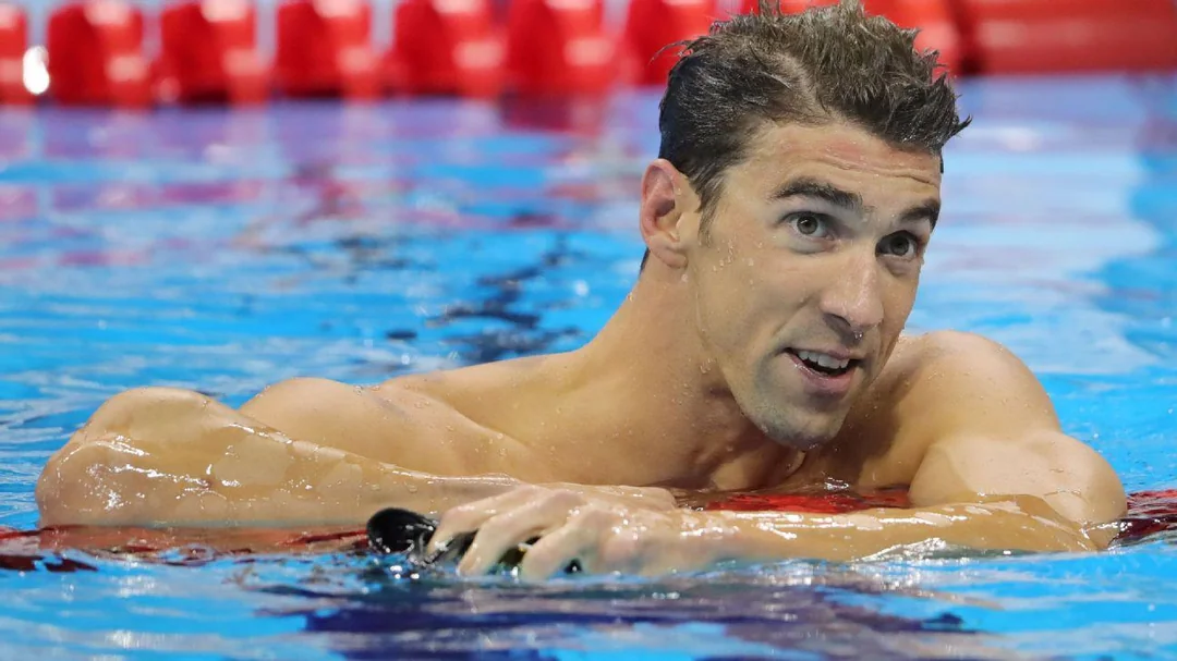 A male swimmer with short dark hair rests at the edge of a swimming pool, holding onto the lane marker, with a background of red lane dividers and water ripples.