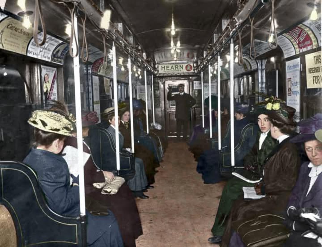 Passengers dressed in early 1900s attire sit inside a vintage subway car, some reading newspapers. The car's interior is lit with overhead lights, and a conductor stands at the far end near the door.
