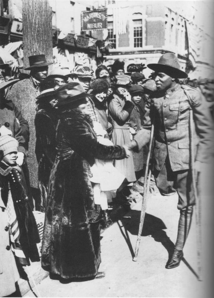 A Black World War I soldier on crutches greets a woman in a crowd during a parade on a city street. Onlookers in winter clothing watch the interaction, with buildings and flags in the background.