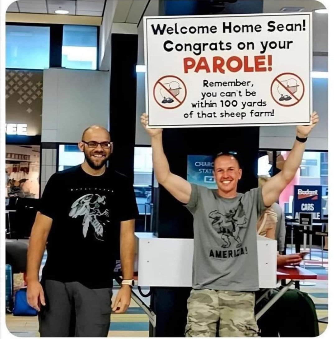 Two smiling men stand in an airport. One holds a large humorous sign that says, “Welcome Home Sean! Congrats on your PAROLE! Remember, you can’t be within 100 yards of that sheep farm!”