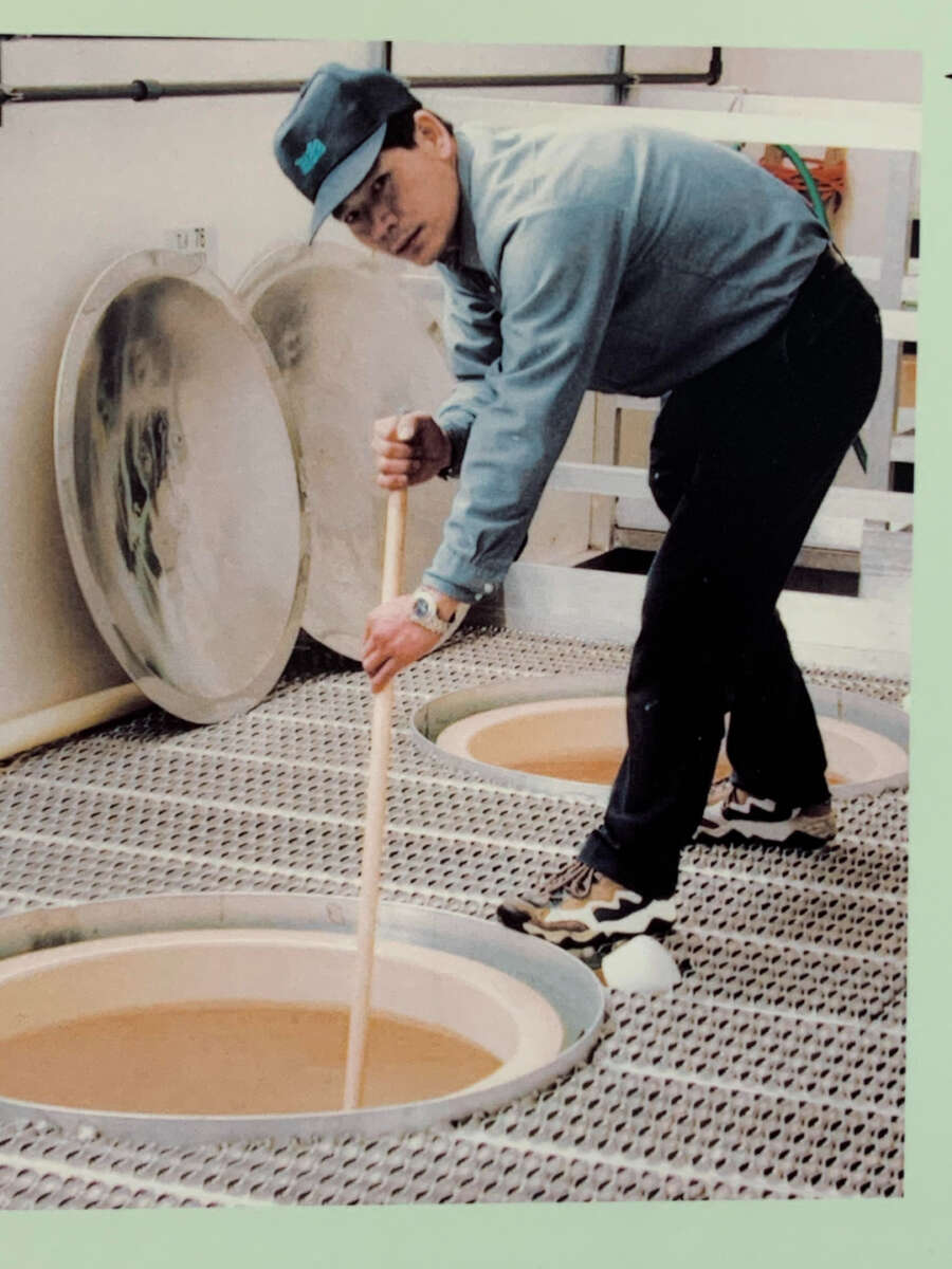A person wearing a cap, glasses, and casual clothes stirs a large vat of brown liquid with a long wooden stick inside an industrial facility. Steam rises from the vat, and there are pipes in the background.
